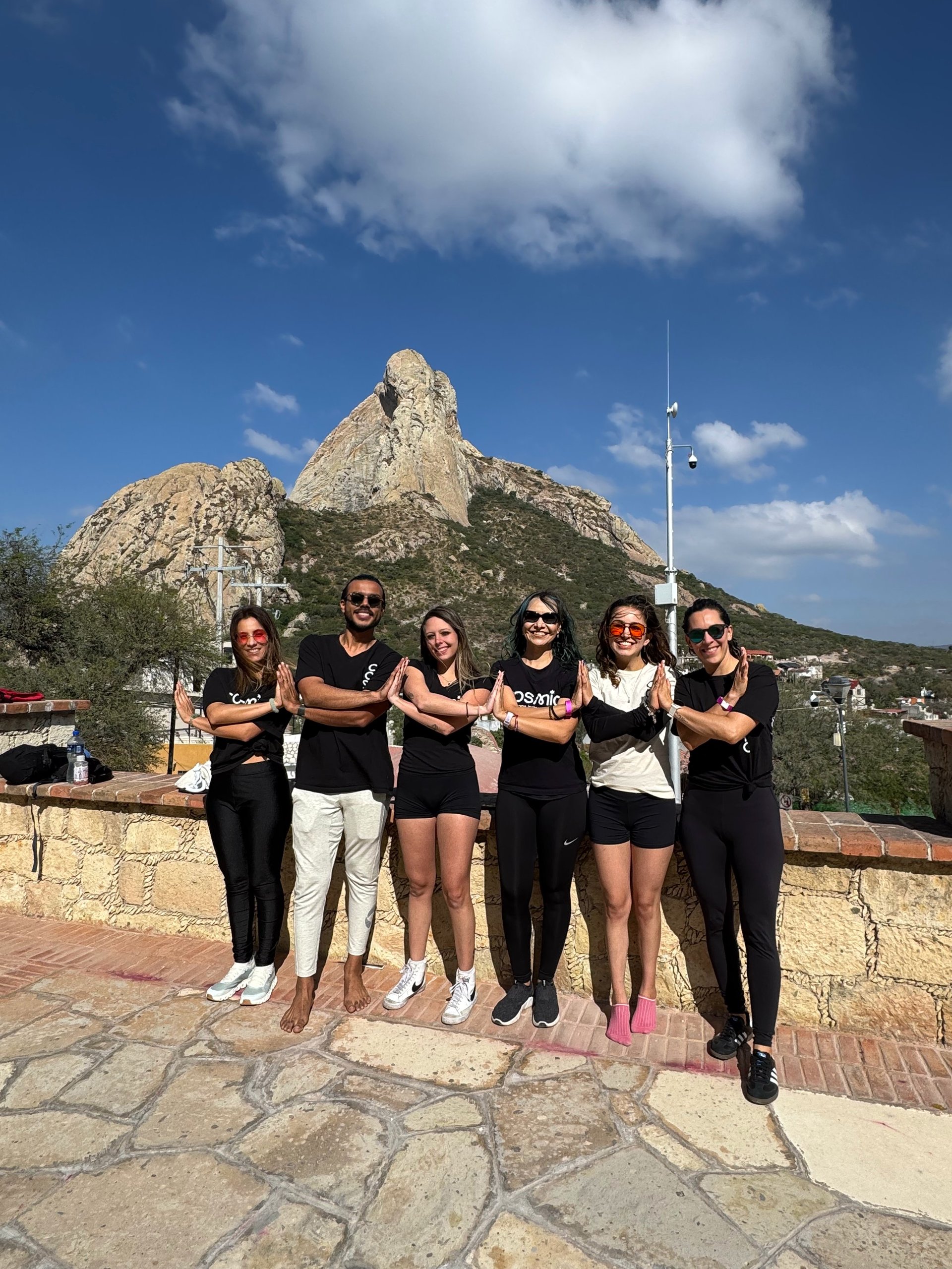 Group of six people standing with arms crossed posing in front of a rocky mountain peak and blue sky