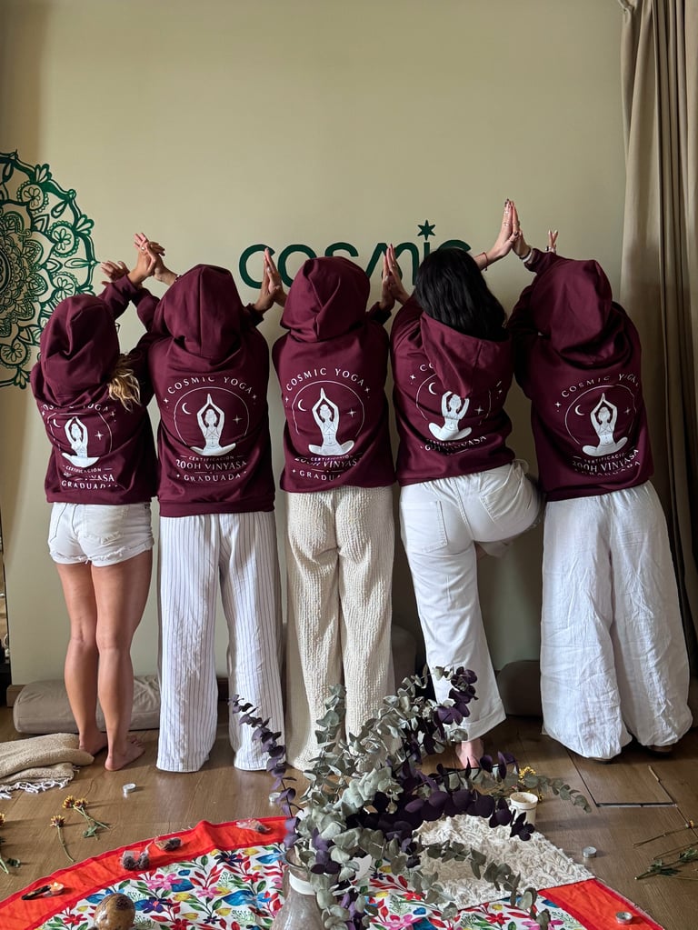 Five people wearing matching maroon yoga hoodies with white pants stand with arms raised, facing away from camera in yoga studio