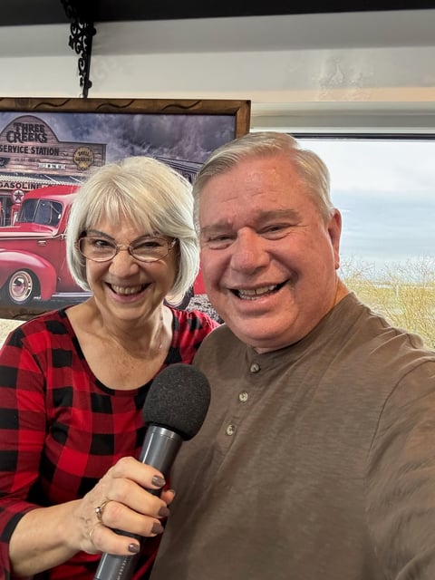 Woman in red plaid shirt and man in brown shirt holding microphone, smiling together indoors with vintage truck poster behind them