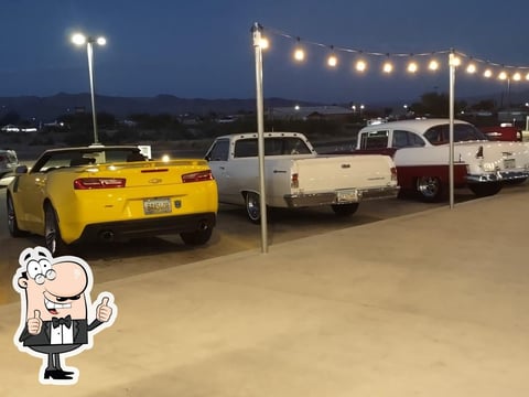Classic and vintage cars parked in a lot at dusk with overhead lighting, including a yellow convertible in the foreground