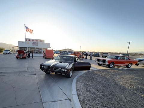 Classic car show at outdoor lot with vintage vehicles, American flag, and spectators under clear sky