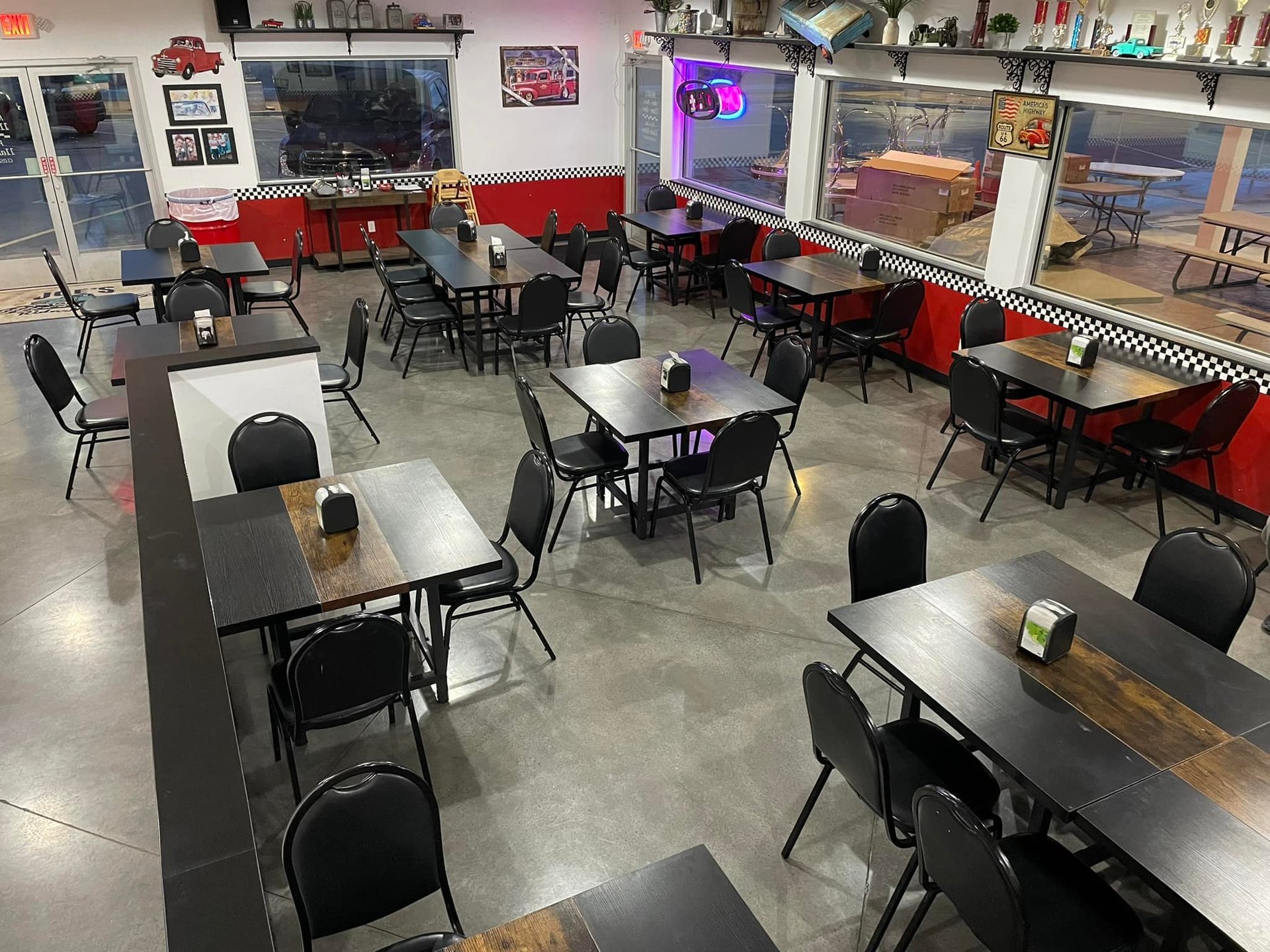 Retro-style diner interior with black tables, red booth seating, black chairs, neon signage, and counter seating along windows.