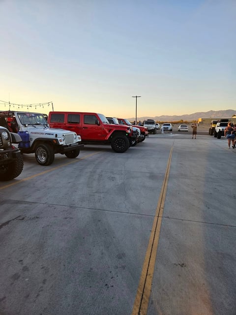 Row of parked vehicles including a white Jeep and red Jeep Wrangler on a desert airfield at sunset with mountains in the distance