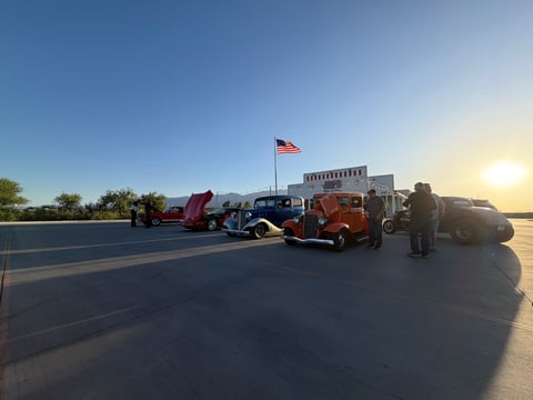 Row of colorful vintage trucks and vehicles parked on a desert road during sunset with an American flag in the background