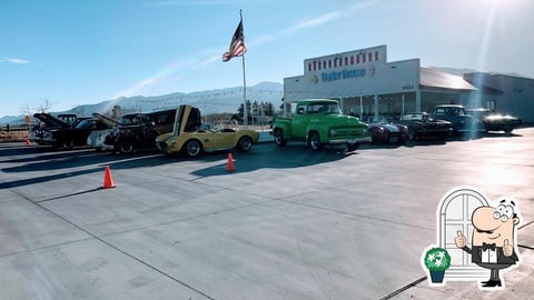 Vintage cars displayed in a lot with mountains in background, American flag, and industrial buildings