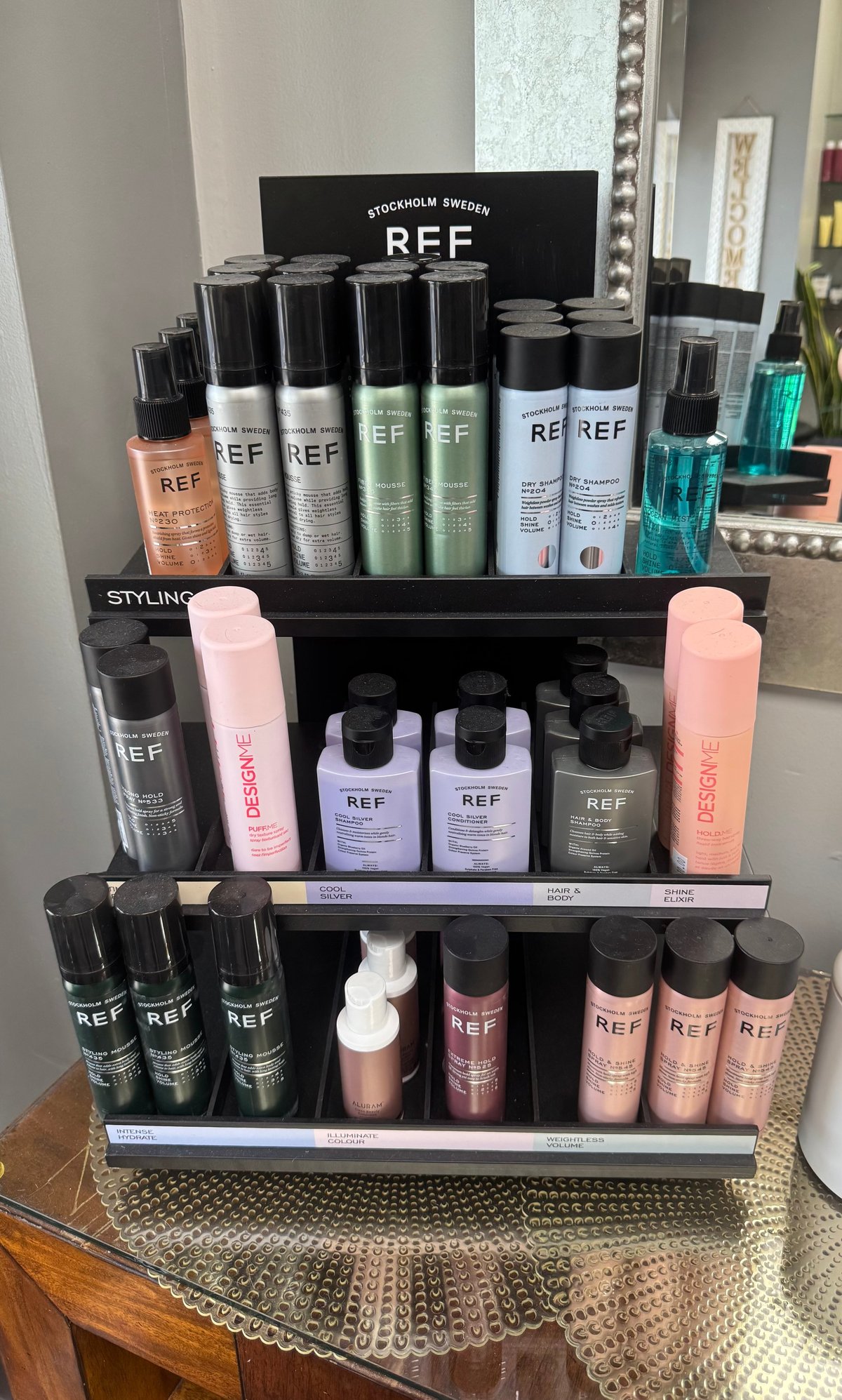 REF hair care product display with multiple shelves of styling and treatment products in black, pink, and clear bottles arranged on a tiered black stand.