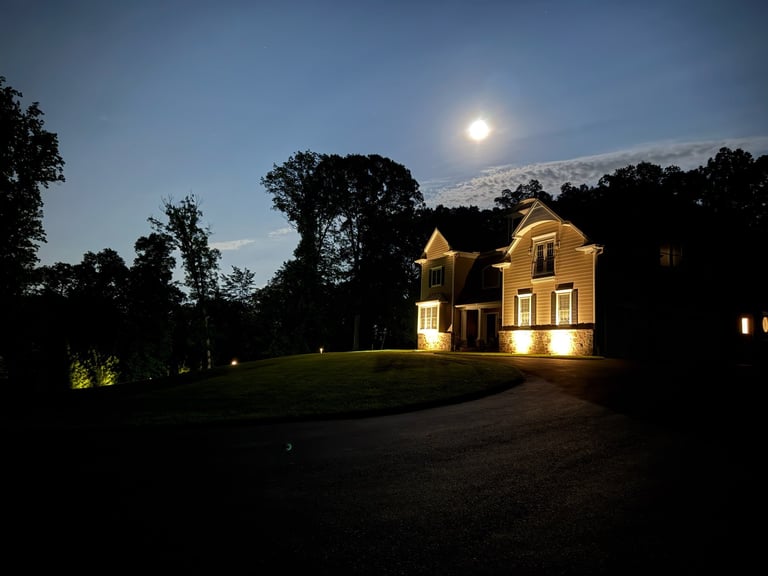 Illuminated yellow house at night with manicured lawn, surrounded by dark trees under a moonlit sky
