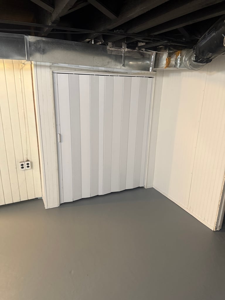 Empty garage interior with white accordion-style door, exposed ceiling ductwork, and electrical outlet on left wall