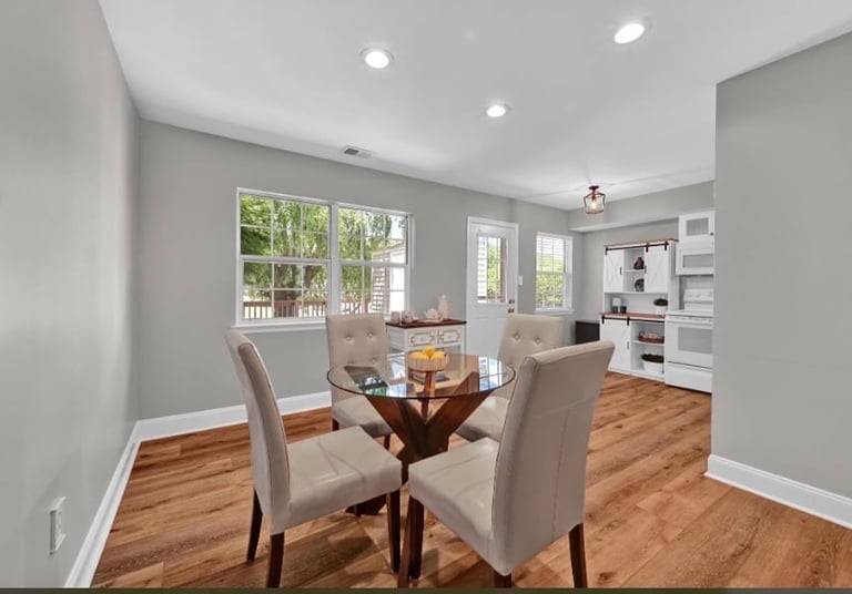 Modern dining room with round glass table, upholstered chairs, hardwood floors, gray walls, and large windows overlooking a deck