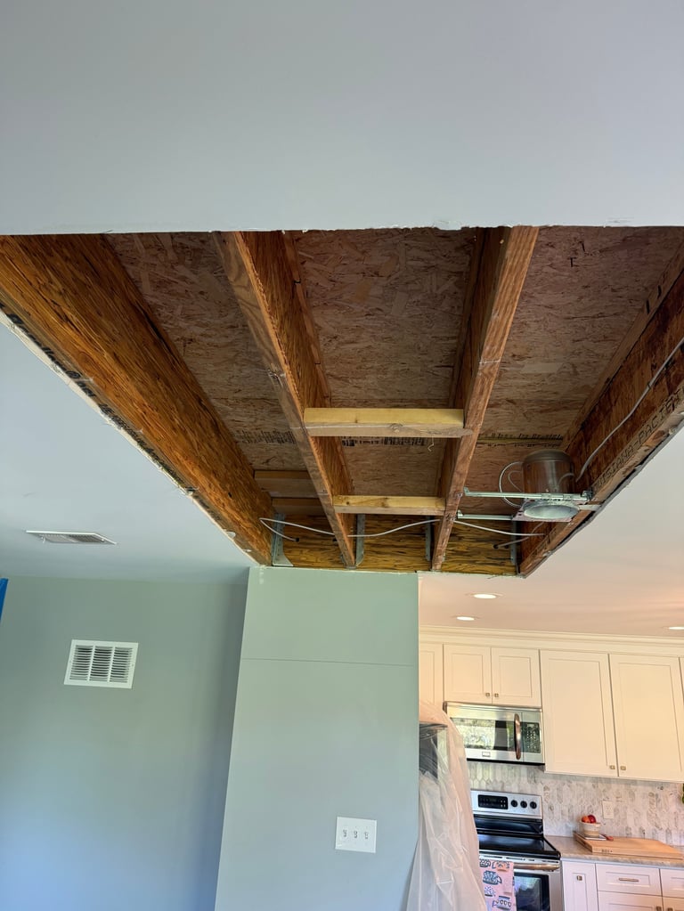 Unfinished ceiling renovation showing exposed wooden beams and joists above kitchen and living room areas with drywall below