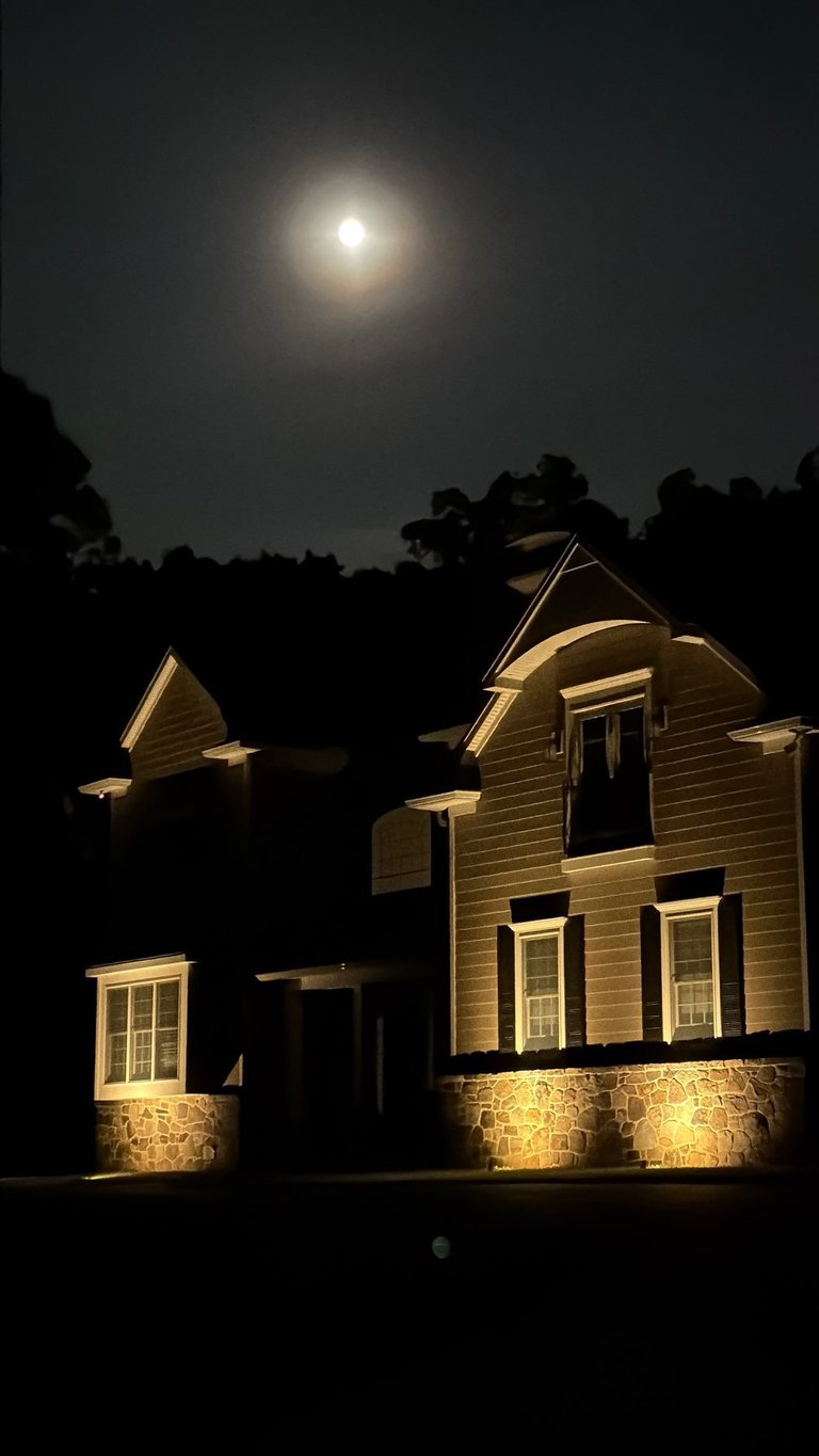 Illuminated colonial-style house at night under a full moon with landscape lighting highlighting the stone foundation and wooden exterior