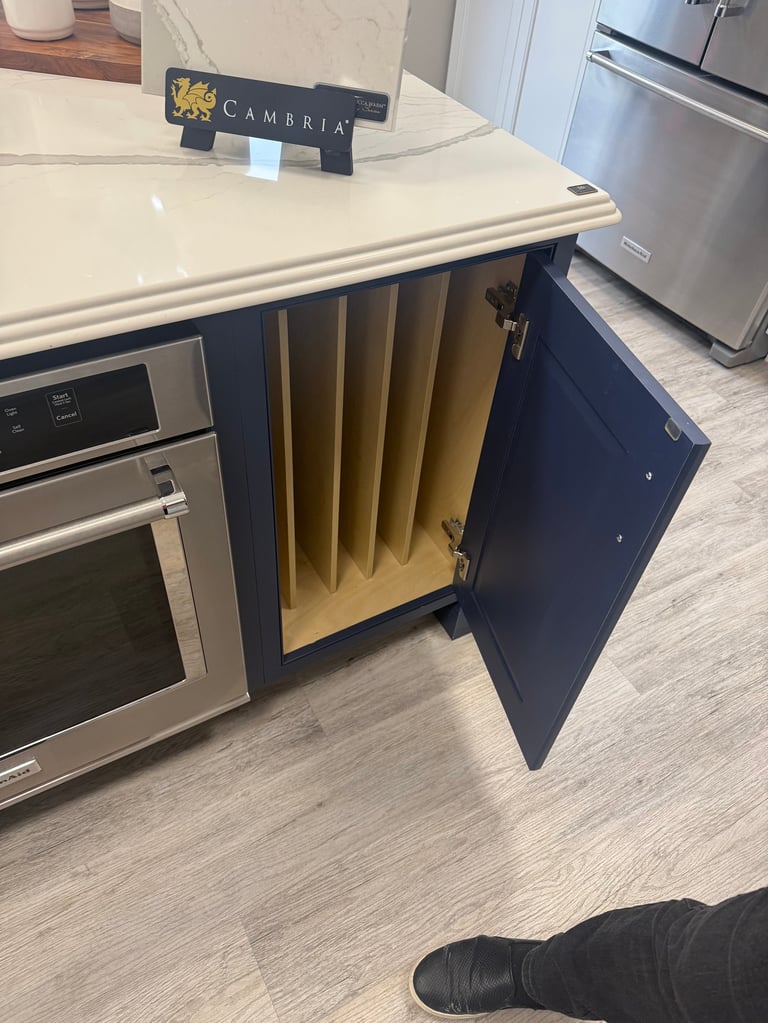 Modern kitchen with cream countertop, dark blue cabinetry with open door revealing gold tray dividers, stainless steel appliances, and Cambria countertop sign