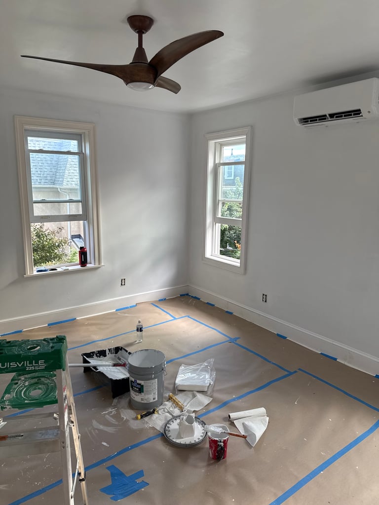 Empty room under renovation with brown ceiling fan, two windows, white walls, blue painters tape on floor, and construction supplies scattered around