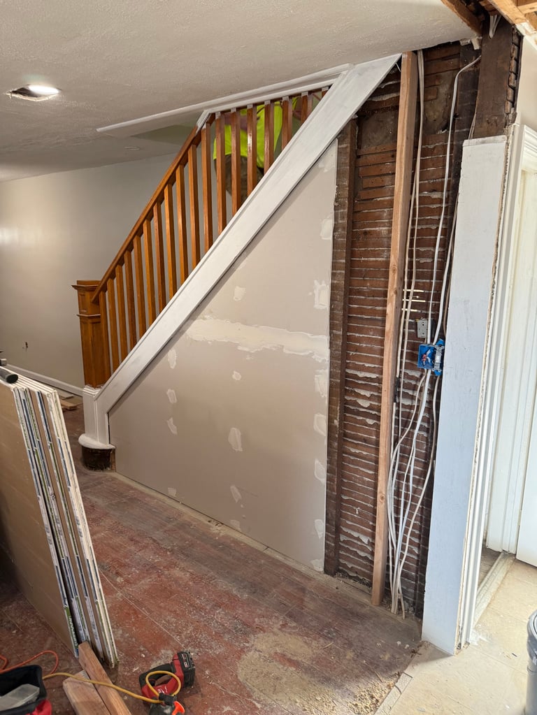 Unfinished basement interior with wooden staircase, drywall patch, exposed brick wall, and construction materials on floor