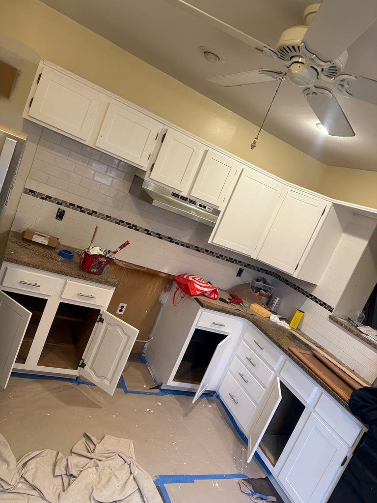 Kitchen under renovation with white cabinets removed, blue painters tape on floor, yellow walls, and exposed countertop areas