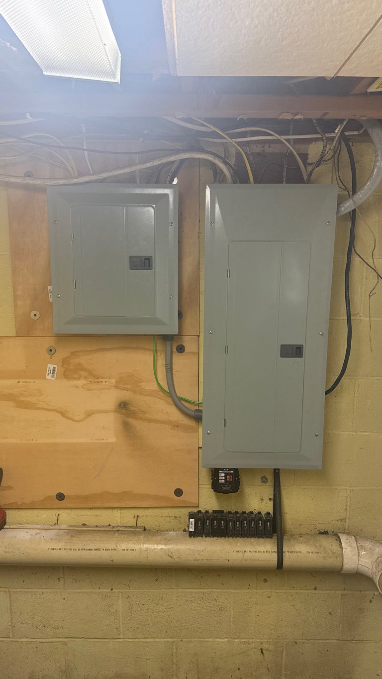 Electrical panel boxes mounted on yellow wall with wiring and conduit overhead in basement or utility space