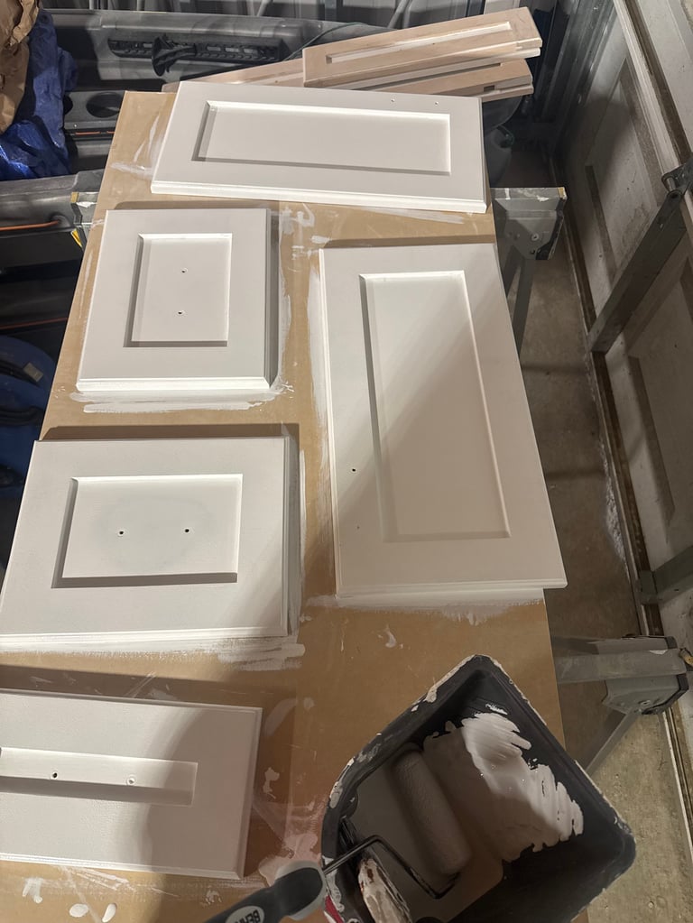 Collection of white cabinet doors and panels arranged on cardboard, various sizes and styles with hardware holes, stored in workshop or storage area