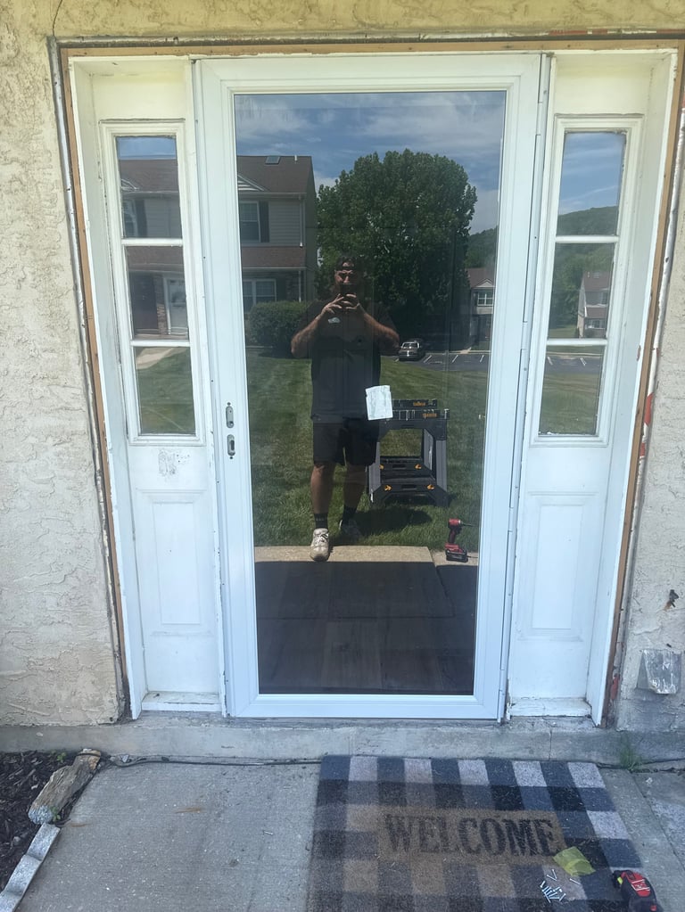 Person in dark clothing taking a selfie reflected in a glass patio door with a welcome mat below
