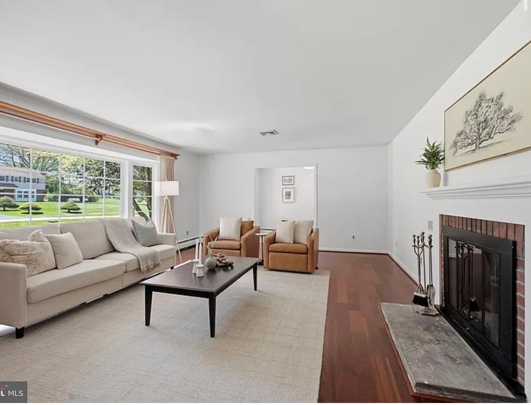 Bright living room with beige sectional sofa, black coffee table, fireplace, hardwood flooring, and large windows overlooking green lawn