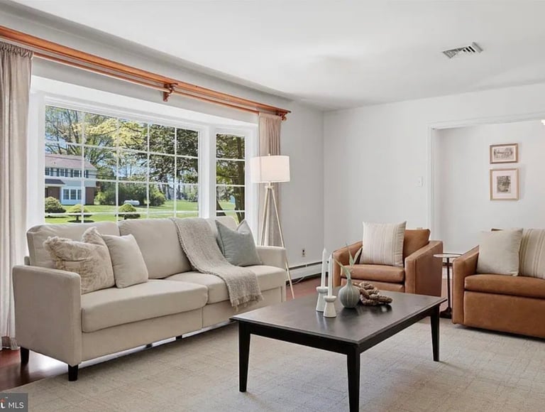 Bright modern living room with beige sectional sofa, cognac leather chairs, black coffee table, and large windows with garden views