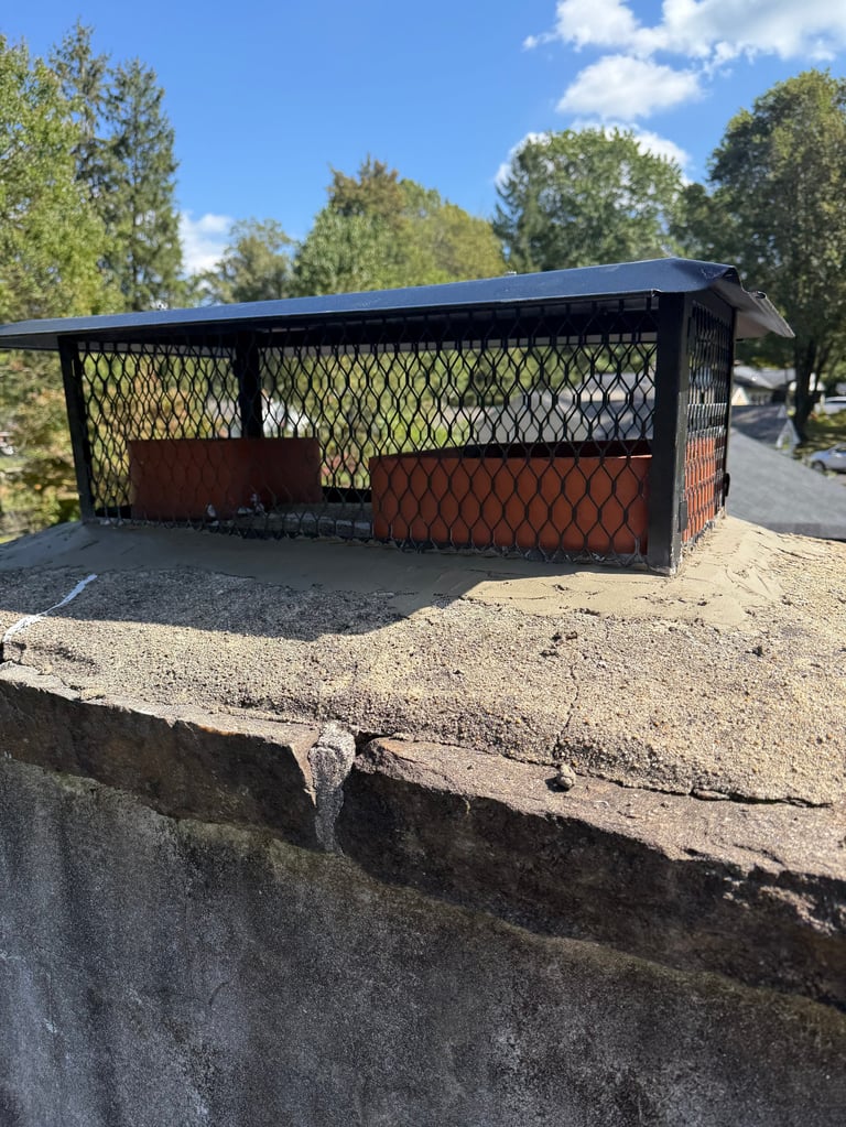 A black metal carport structure with mesh panels and rust-colored backing, situated on a gravel lot with trees in the background under blue sky