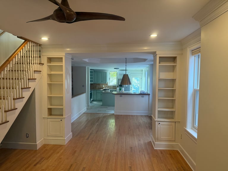 Modern open-concept home interior with hardwood floors, cream-colored built-in shelving, staircase on left, kitchen visible through archway, and pendant lighting