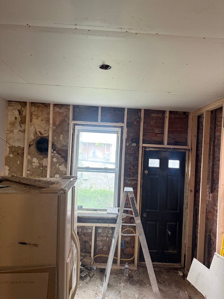 Home kitchen renovation in progress showing exposed walls, drywall, a step ladder, window with black door, and appliances