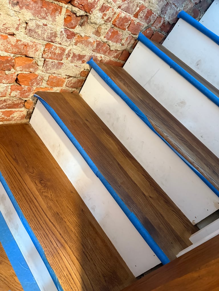 Wooden floor samples with blue tape against exposed red brick wall, displaying different wood finishes and colors