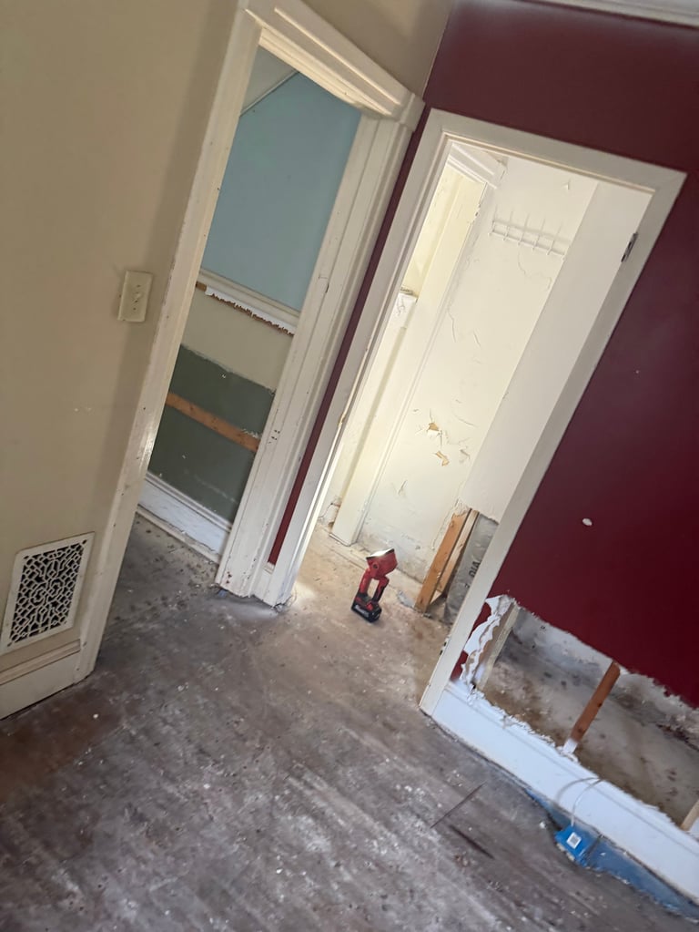 Interior room under renovation with exposed subfloor, white trim around doorways, maroon and tan walls, and construction materials scattered on the floor