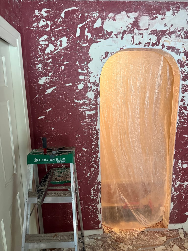 A large wooden door frame wrapped in plastic sheeting on a weathered burgundy and white wall with a green Louisville ladder beside it