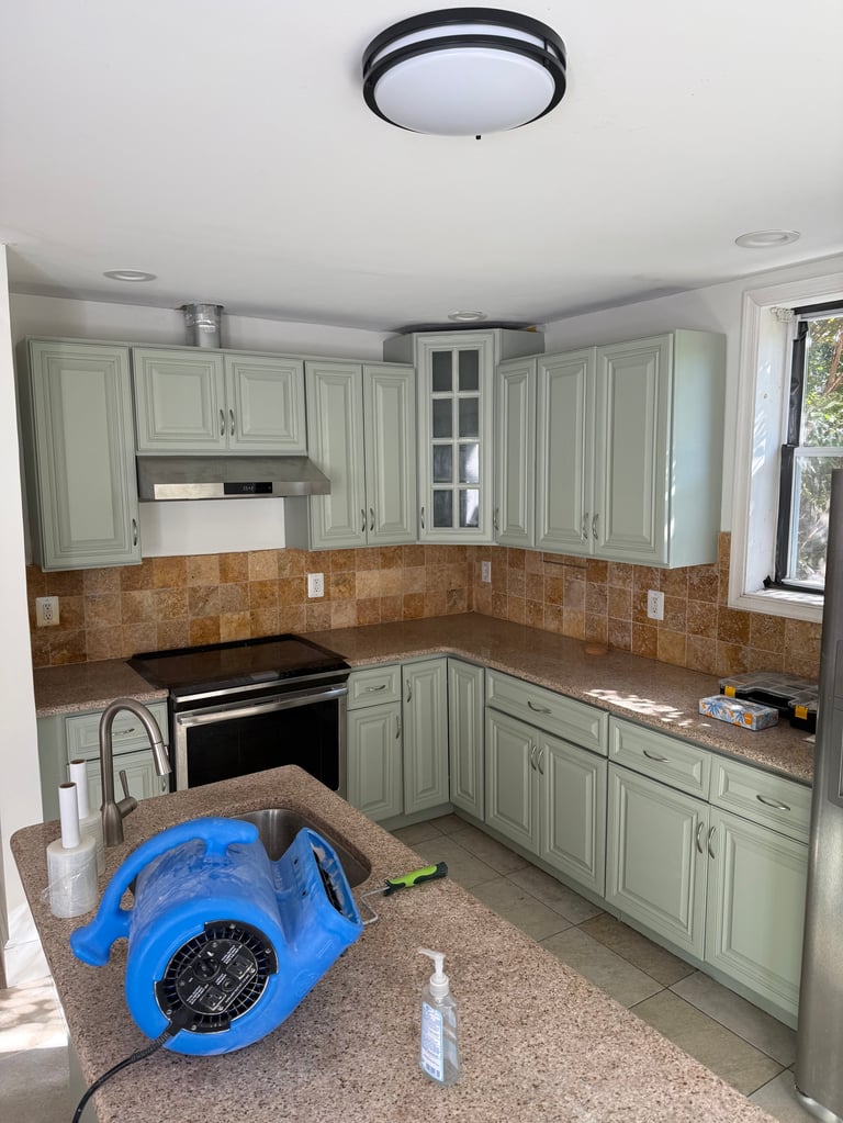 Kitchen with light green cabinets, brown tile backsplash, and blue drying fan on the counter during renovation