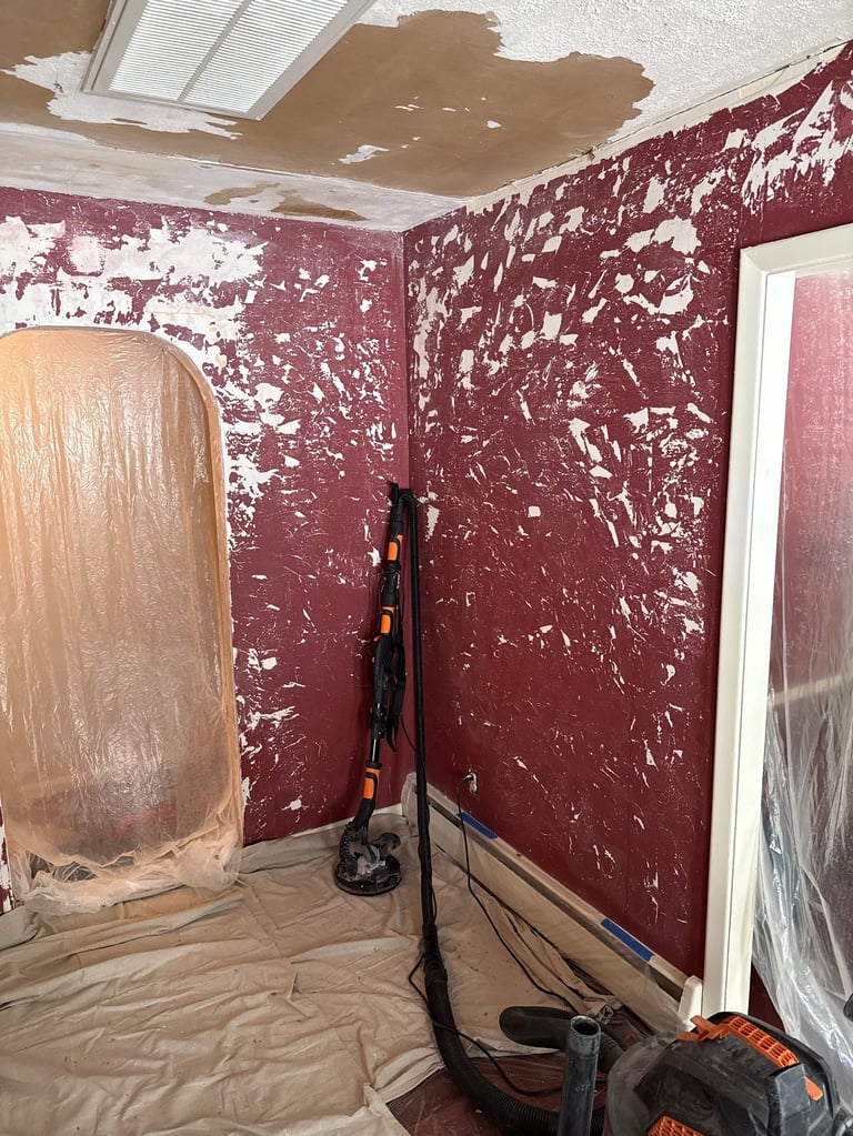 Interior room under renovation with burgundy walls showing peeling paint, damaged ceiling drywall, and construction equipment on plastic-covered floor