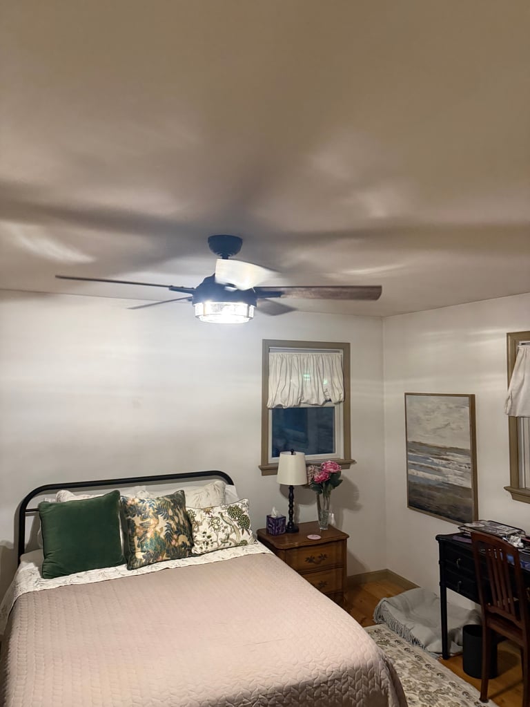 Bedroom with blue ceiling fan, bed with green pillows, wooden nightstand with flowers, and framed artwork on walls