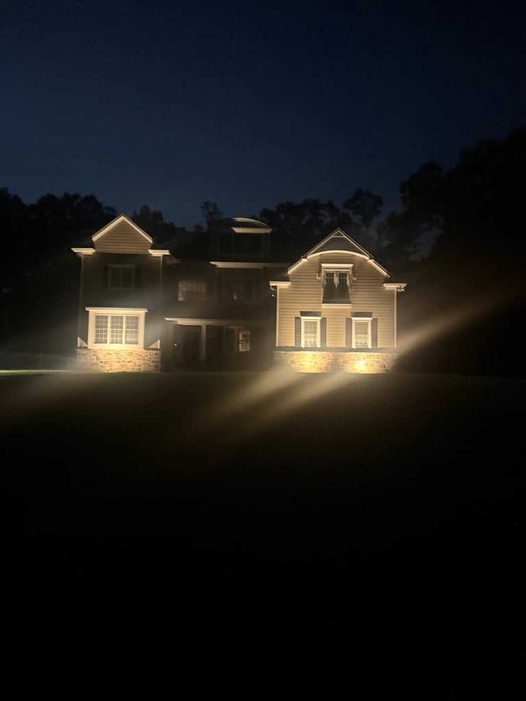 Two illuminated colonial-style houses at night with warm exterior lighting, surrounded by dark trees against a twilight sky
