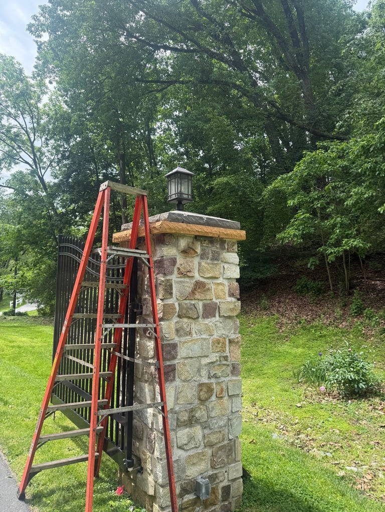 Red ladder leaning against a stone pillar with a lamp on top in a green yard surrounded by trees