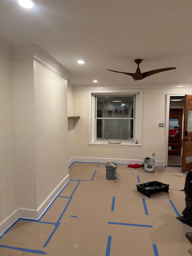 Room under renovation with blue painters tape on the floor, beige walls, ceiling fan, and window