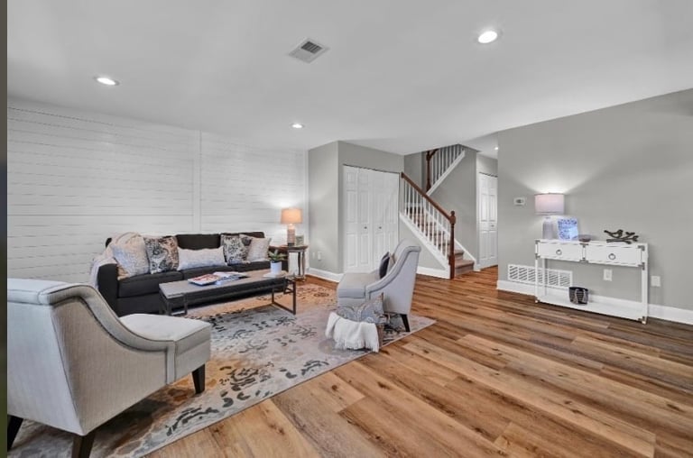Modern living room with hardwood floors, gray walls, black and beige seating, white fireplace, wooden staircase, and recessed ceiling lights