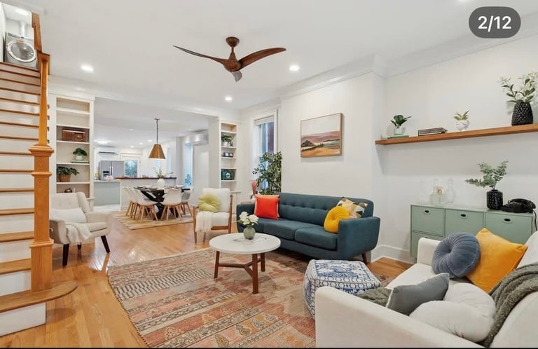 Bright modern living room with blue sofa, armchairs, ceiling fan, wooden shelving, and open kitchen layout