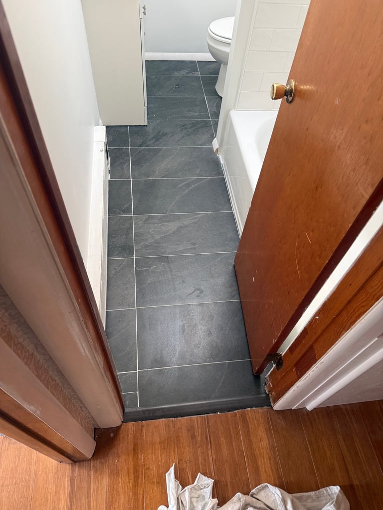 View into a narrow bathroom with dark slate tile flooring, white walls, wooden doors, and a toilet visible in the background