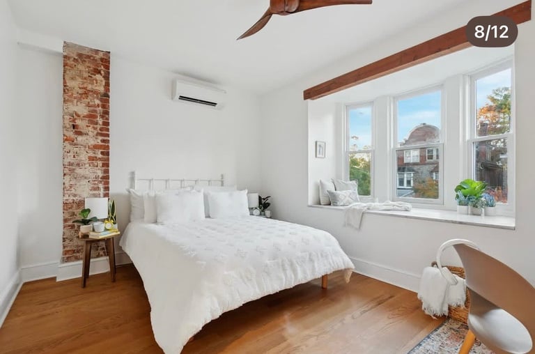 Bright modern bedroom with white bed, exposed brick accent wall, wooden beams, hardwood floors, and large windows overlooking street