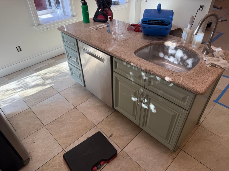 Kitchen island with brown granite countertop, sink, and sage green cabinetry in a tiled kitchen with various items on counter