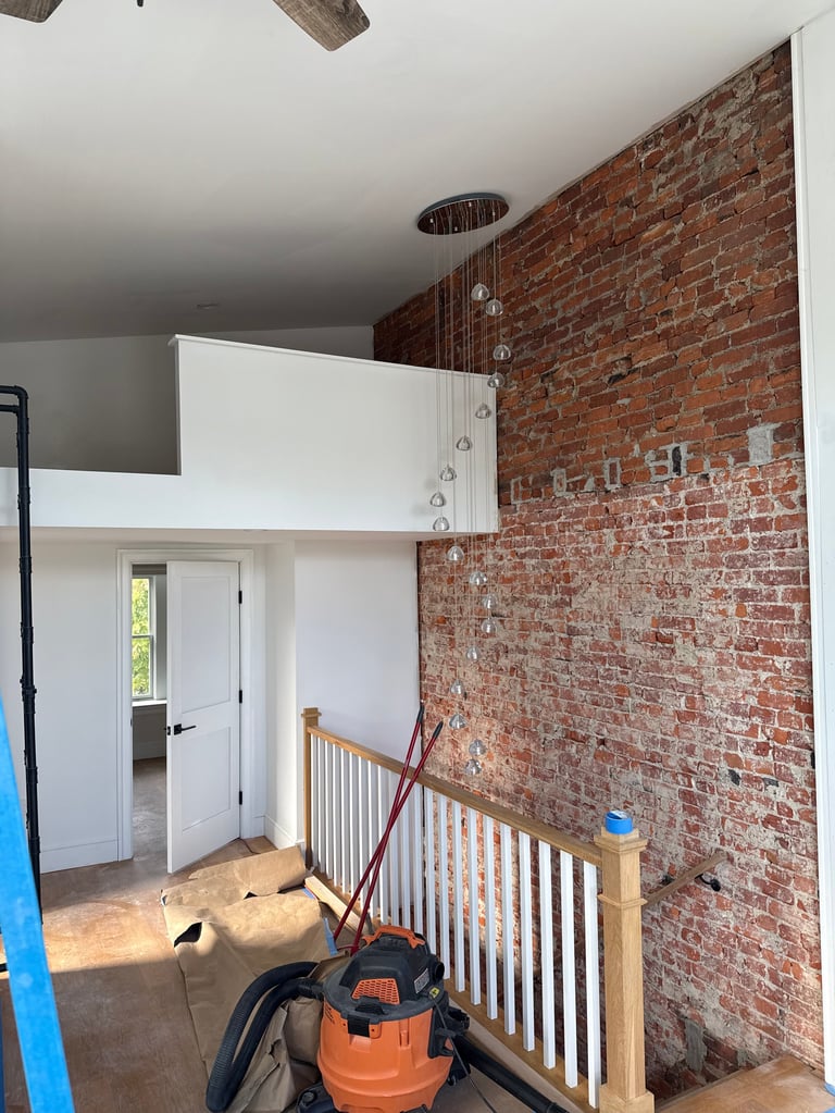 Modern loft bedroom with white platform bed, exposed red brick wall, wooden railing, and construction equipment on floor