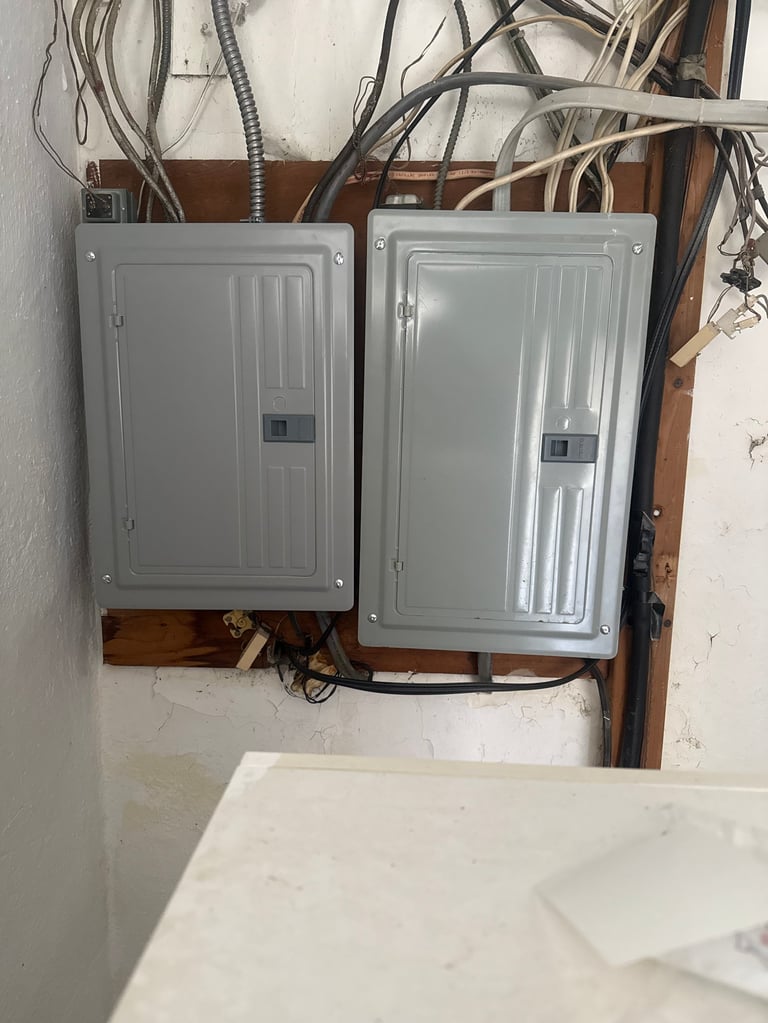 Two gray electrical circuit breaker panels mounted on a wooden board with tangled wires above them on a white wall