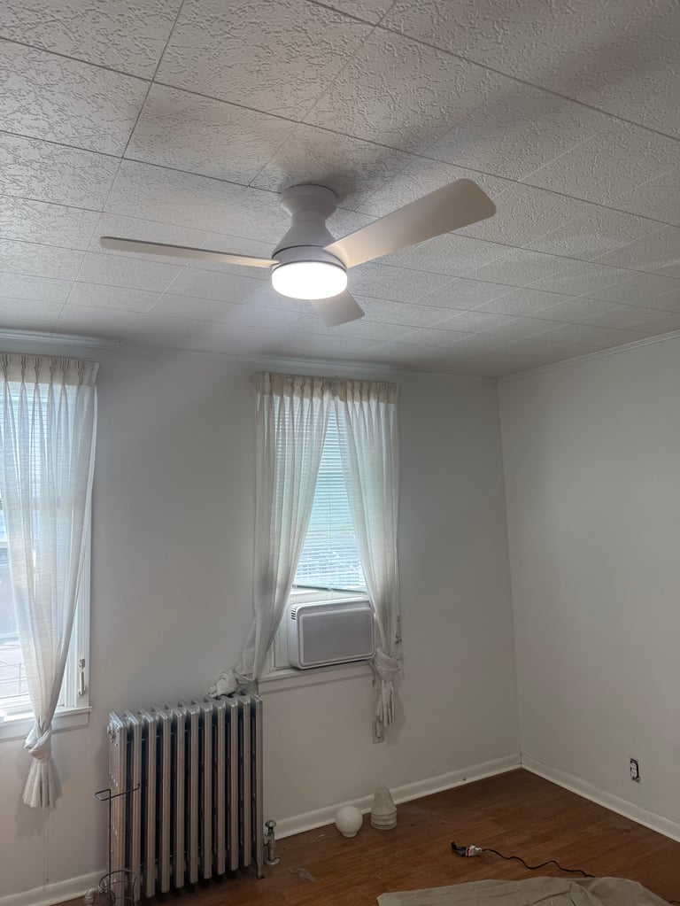 Modern ceiling fan with LED light mounted in empty room with white walls, textured ceiling, two windows with curtains, and hardwood floor
