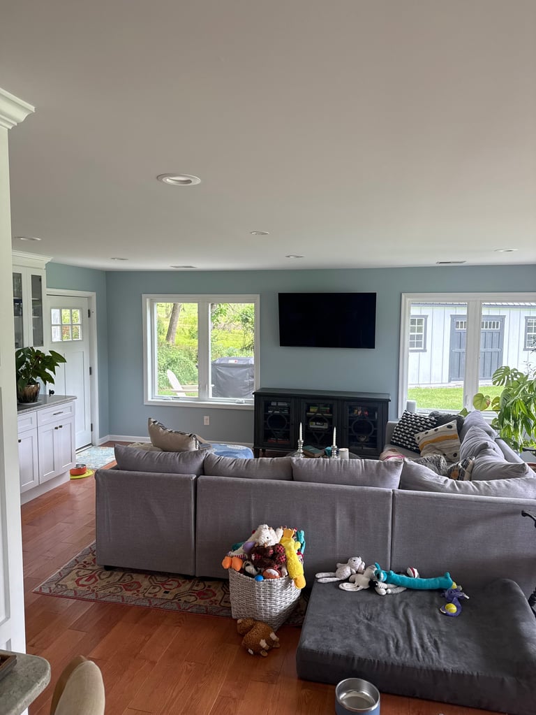 Modern living room with gray sectional sofa, TV above black entertainment center, hardwood floors, and large windows overlooking green yard