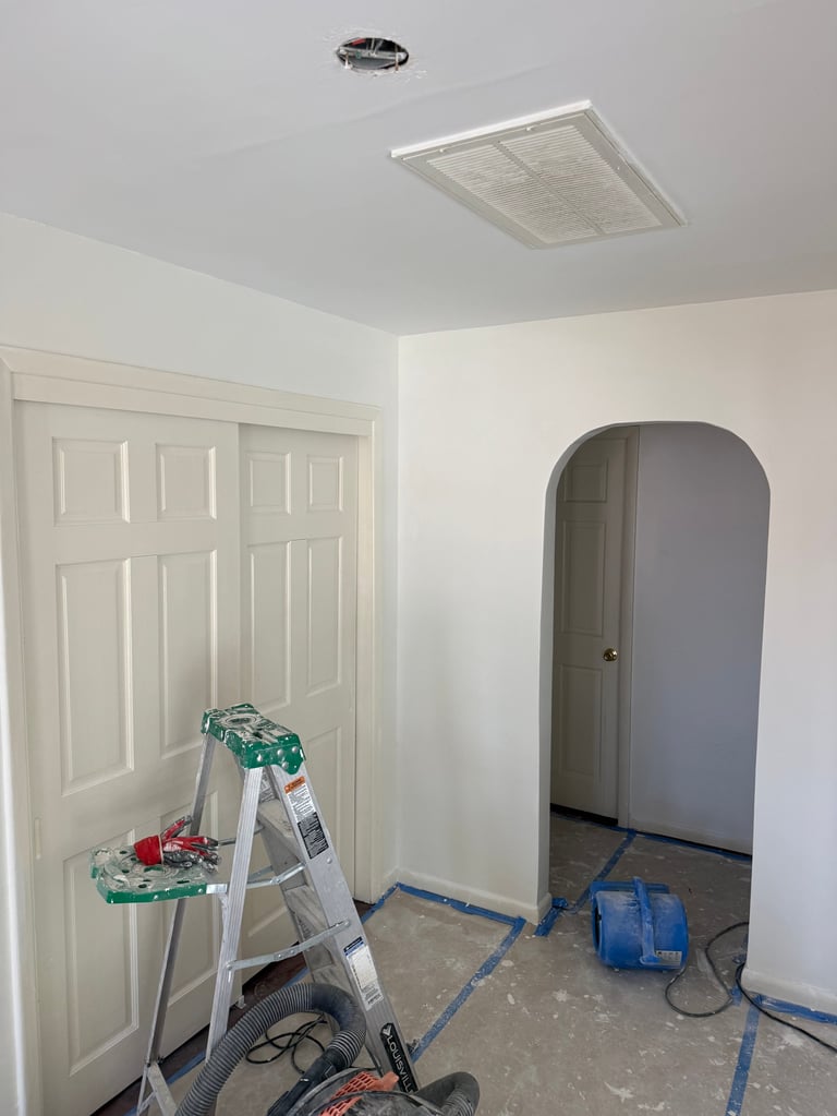 Interior room under construction with white double doors, arched doorway, ladder with tools, ceiling light fixture, and blue plastic sheeting on floor