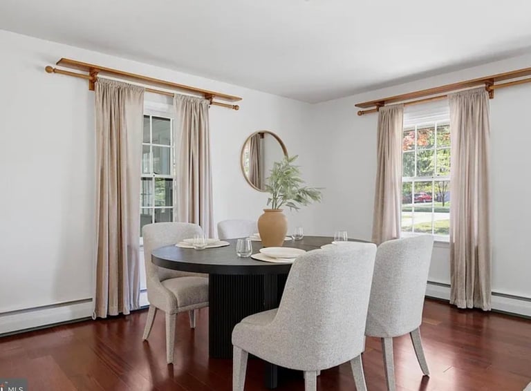 Modern dining room with black round table, upholstered cream chairs, wooden floors, and floor-to-ceiling windows with draped curtains