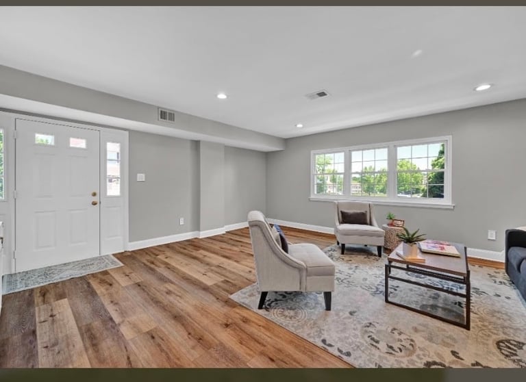 Bright modern living room with hardwood floors, gray walls, white ceiling with recessed lighting, large windows, and minimal furniture including armchair and media console