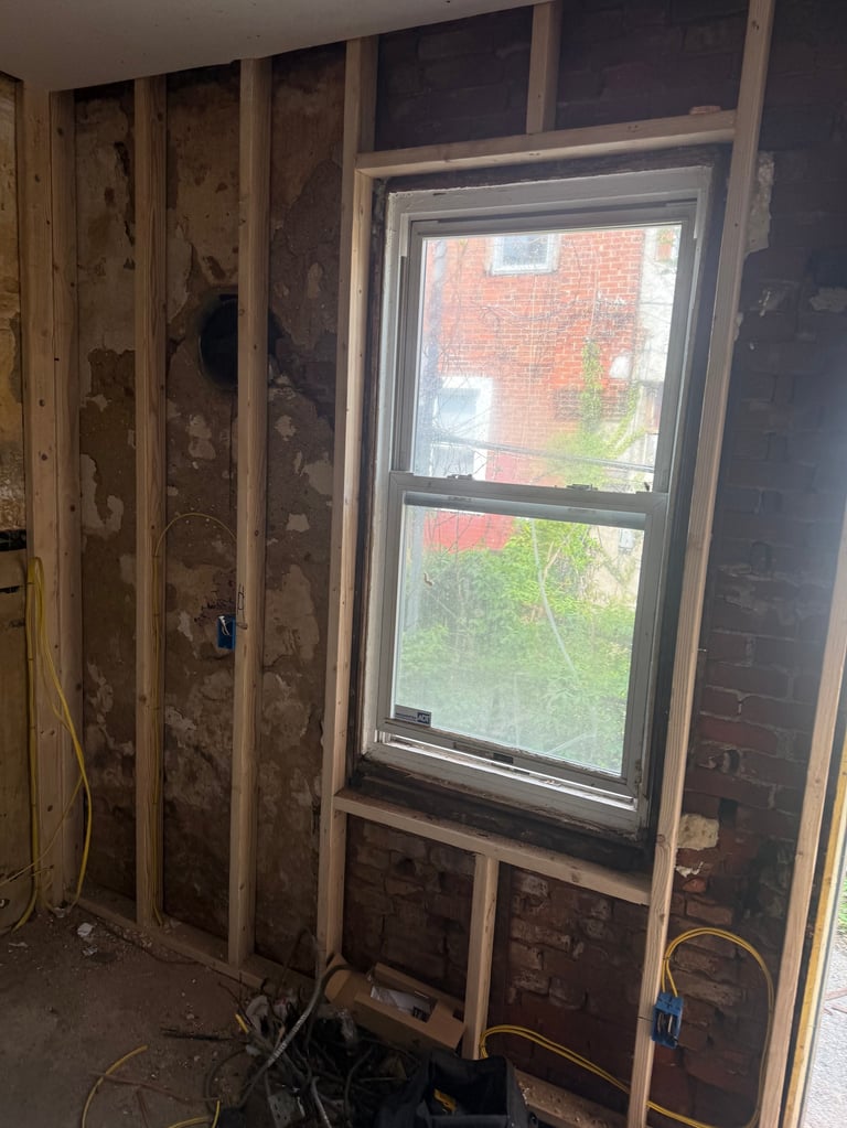 Interior wall framing with wooden studs and exposed insulation next to a double-hung window overlooking a brick building and yard
