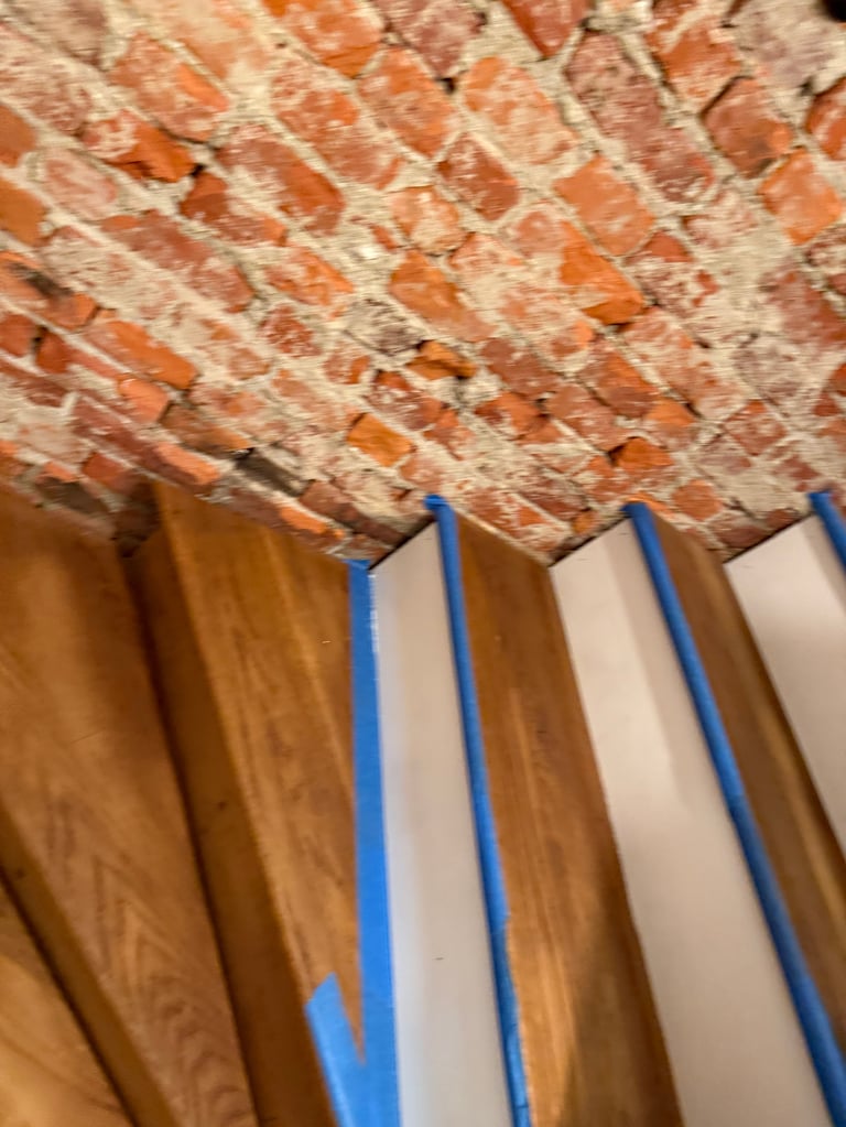 Blue painters tape applied to wooden surface with exposed brick wall above, indicating wall or trim preparation for painting